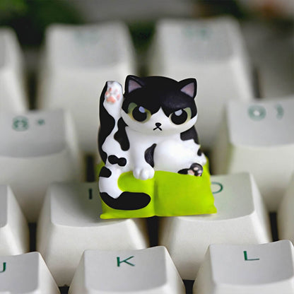 Keycapper Cat Grooming artisan keycap featuring a cute black and white Tuxedo cat lifting its leg on a lime green cushion.