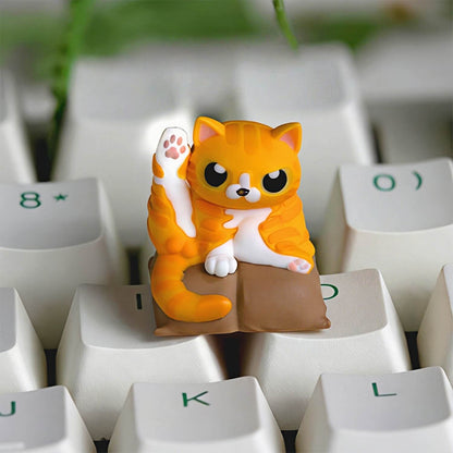 Keycapper Cat Grooming artisan keycap featuring a chubby orange tabby cat doing a leg stretch pose on a brown cushion.